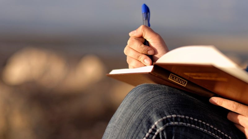 A person writes with a blue in a brown journal book with with it propped on their knee while sitting outside.