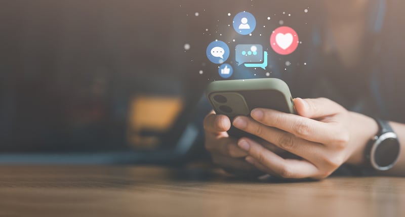 Hands hold a smartphone with icons representing social media are above it.
