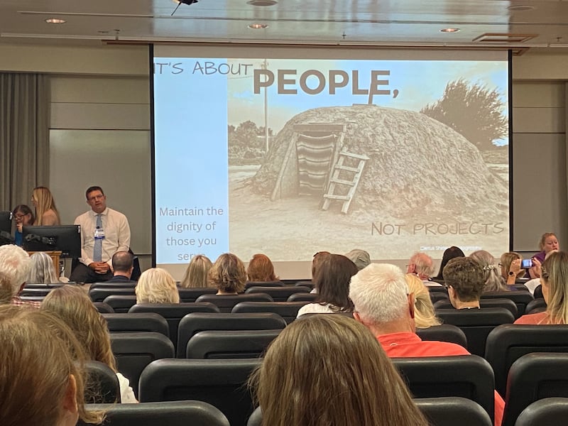 Chris Crippen&nbsp;sitting at the front of a lecture hall, next to a presentation slide that says “It’s about people, not projects” with a picture of a hogan.