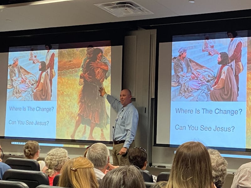 Gregory Johnson&nbsp;at the front of a lecture hall, standing next to a presentation slide with an image of Peter healing a paralytic and an image of the prodigal son hugging his father.