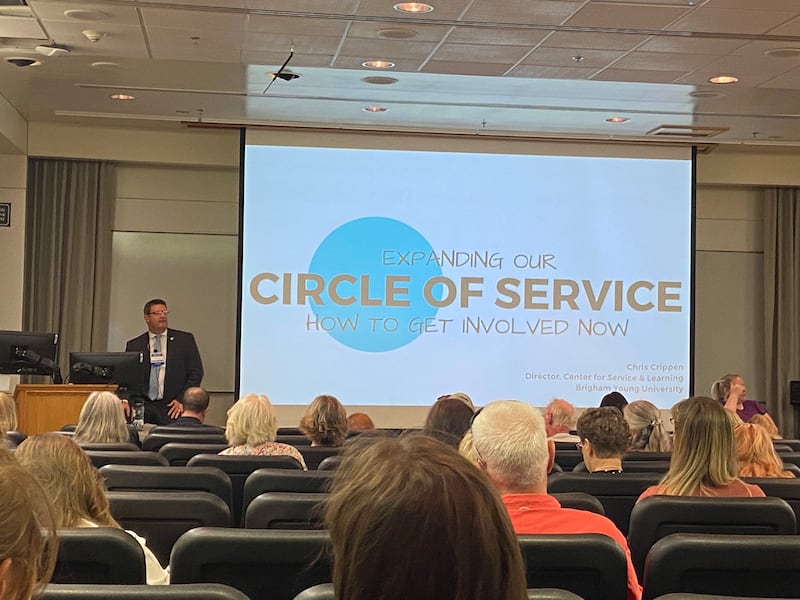 Chris Crippen&nbsp;sitting next to a presentation slide that says “Expanding Our Circle of Service” at the front of a lecture hall.