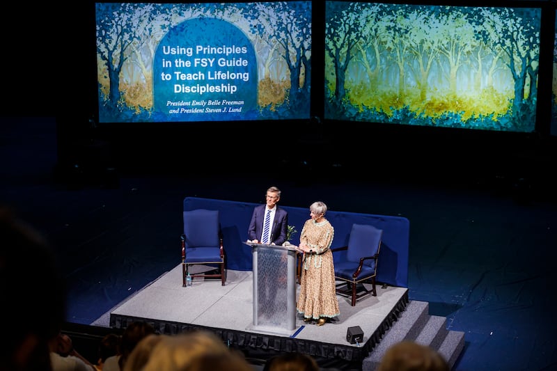 President Emily Belle Freeman and President Steven J. Lund stand at the podium in the Marriott Center with a screen behind them saying "Using Principles in the FSY Guide to Teach Lifelong Discipleship."