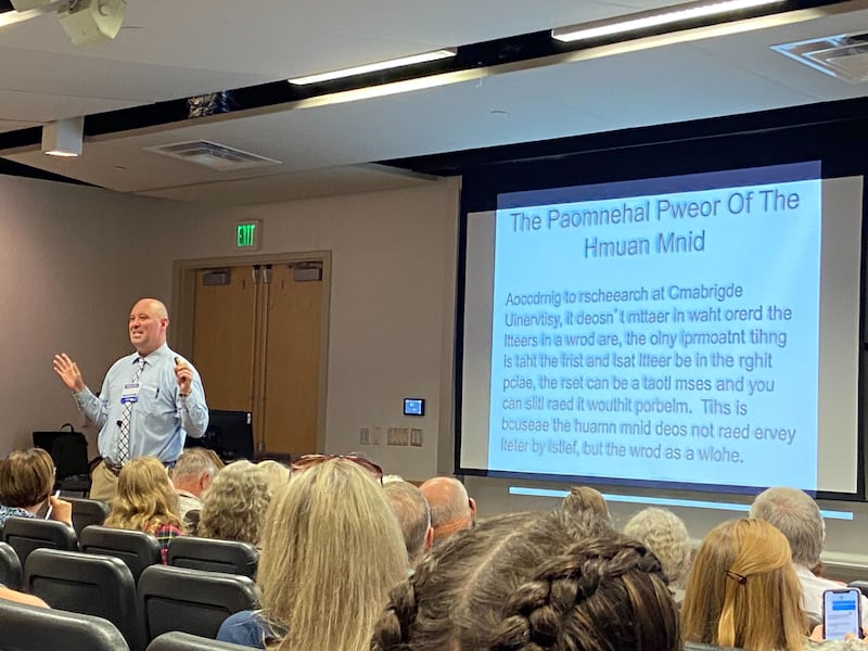 Gregory Johnson&nbsp;at the front of a lecture hall, standing next to a presentation slide that says “The Phenomenal Power of the Human Mind” with only the first and last letter of each word in the correct place.